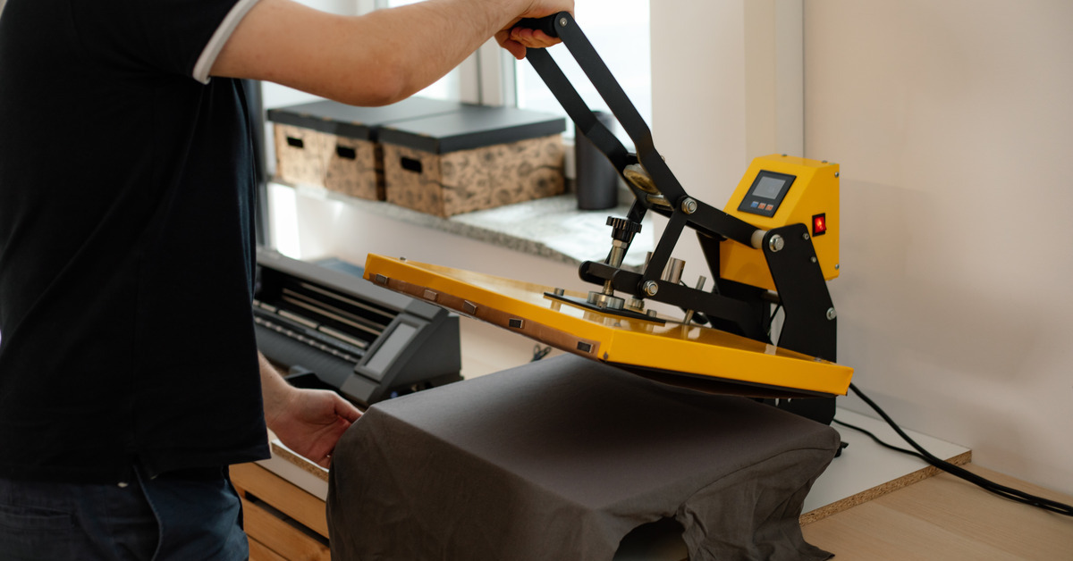 A man in a black T-shirt grabbing the handle of a T-shirt printing press. On the press is a dark gray shirt with no design on it.