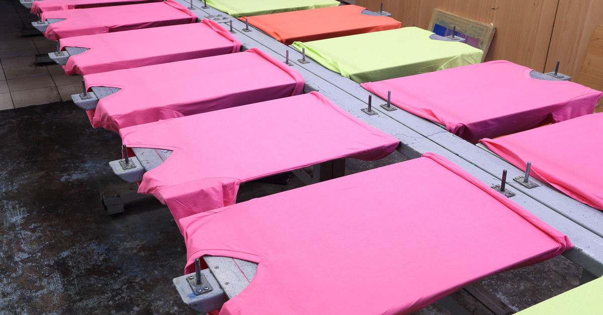 Several blank pink T-shirts spread out on a screen table with their front sides facing up. In the background are a few white shirts.