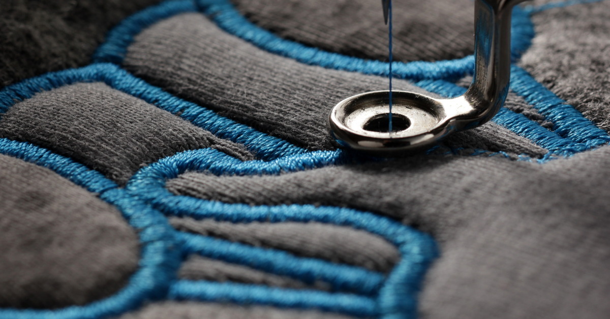 Closeup of a gray piece of clothing with a detailed embroidered blue pattern on it. A sewing machine is guiding the thread.