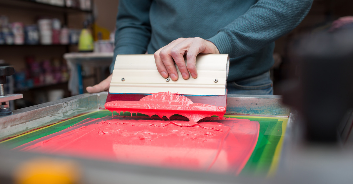 Close-up of a screen printing frame. A man is using a tool to spread a large amount of bright pink color.