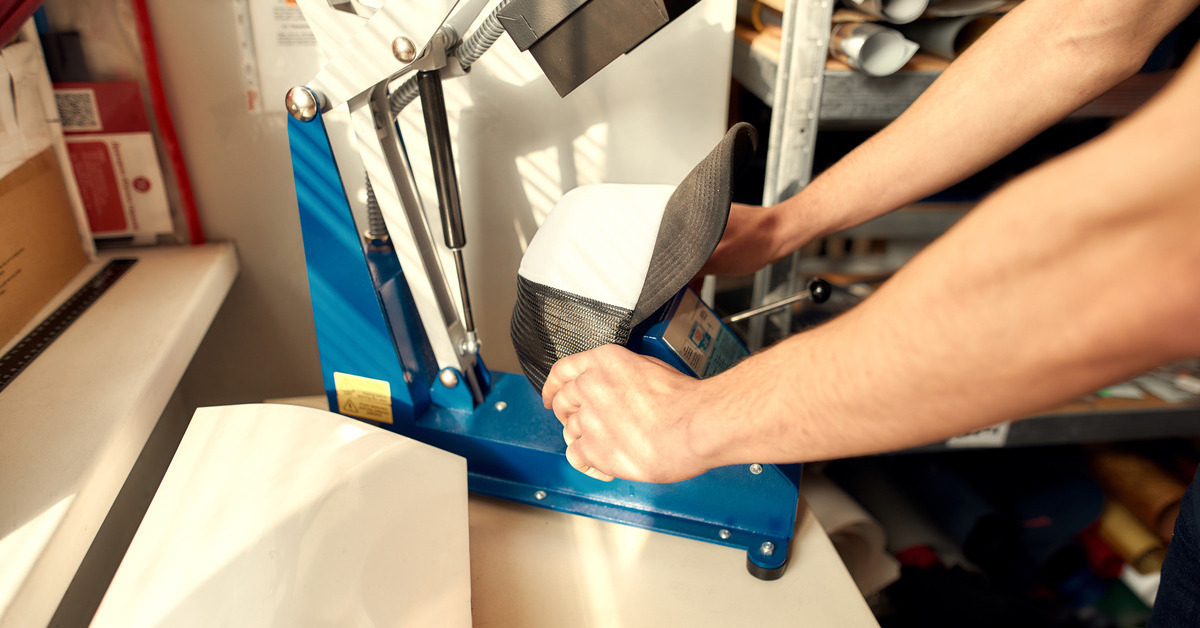 Pair of hands loading a black and white baseball cap into a printing press machine. The front canvas of the hat is blank.