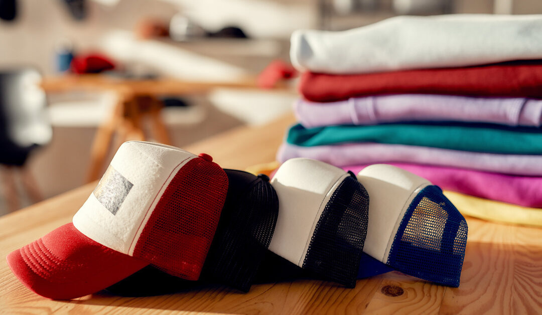 Four baseball caps of differing colors are linked together on a wooden table. Behind them is a pile of folded sweaters.