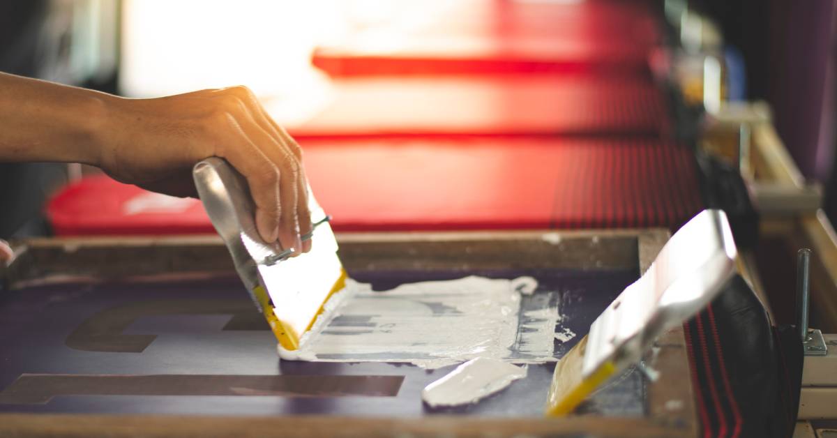 T-shirt printing process. A pair of hands using a frame and ink station to rub white ink onto a red t-shirt.