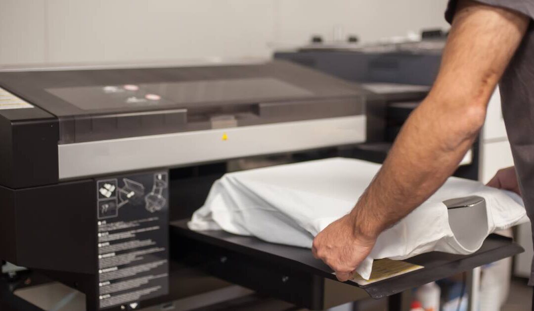 Man in gray shirt is carefully folding and loading a blank white t-shirt into a large printing machine.