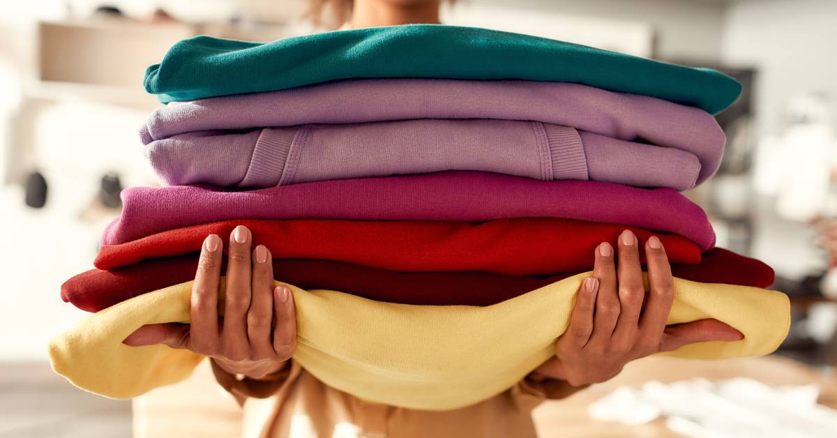 A person smiles while holding a stack of sweatshirts. Each one is a different color, including reds, purples, yellow, and cyan.