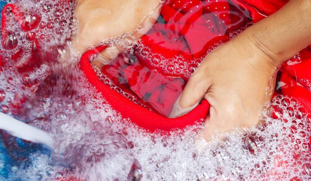 A woman handwashing a red polo shirt with white spots in a blue tub. She uses both hands, dipping the shirt into soapy water.