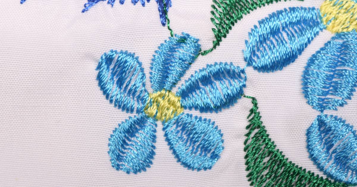 White fabric with a detailed, embroidered pattern on it. The pattern contains blue flowers and green leaves.